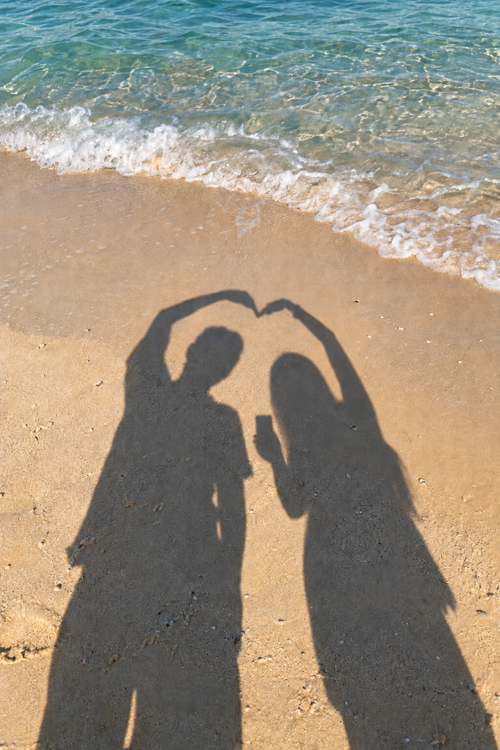 きれいな海の浜辺で、二人の影がハートを作り、女性が左手のスマホで撮影している写真